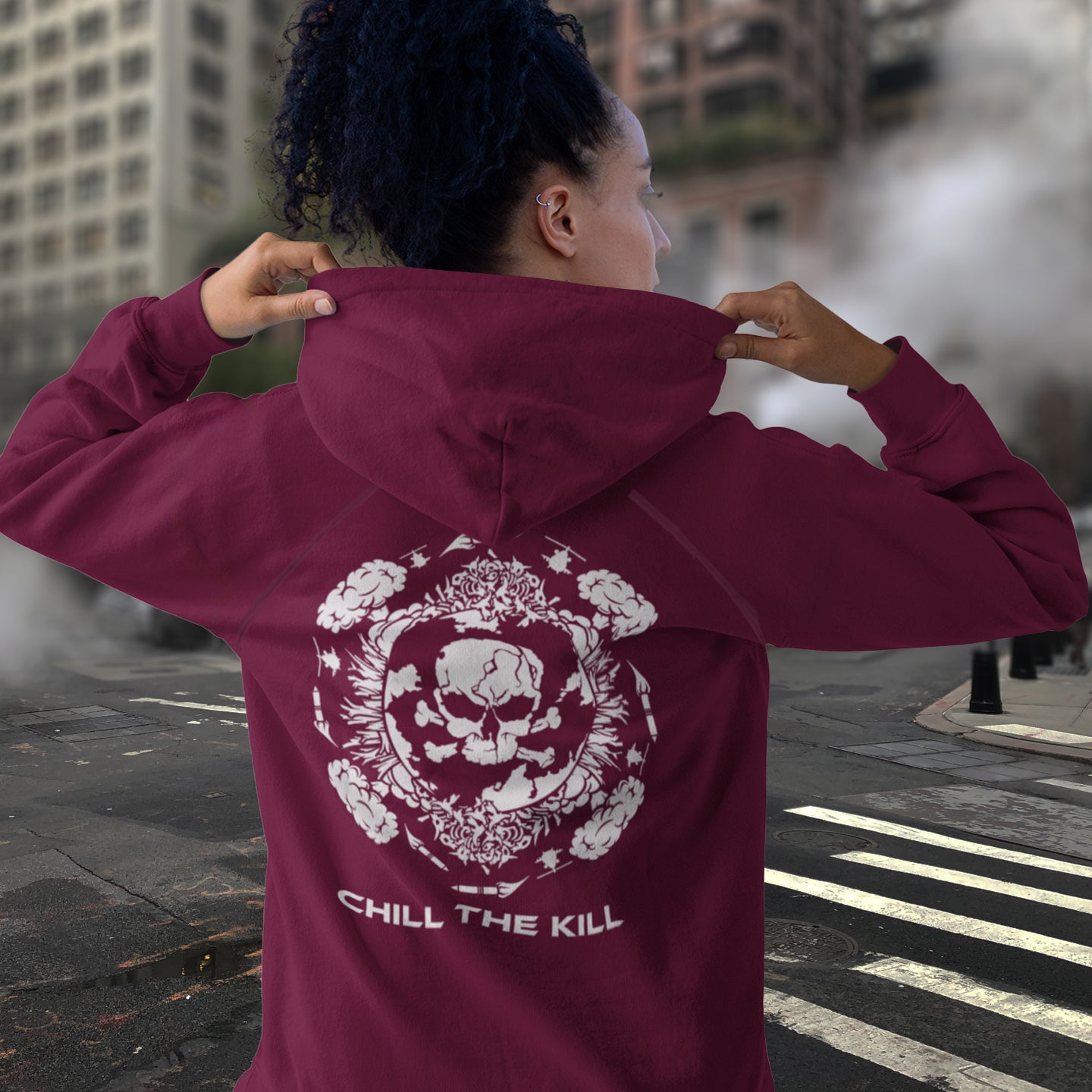 Person wearing a burgundy hoodie with a skull design and 'Chill the Kill' text, standing on a city street. A 'save the planet' hoodie about human impact on nature. An Earth day special. This design highlights the amount of death and destruction we create as a species. Comfortable and practical. It's extra soft and has a convenient front pouch pocket. Made from organic cotton and recycled polyester, this anti-war political hoodie is a great eco-friendly choice.