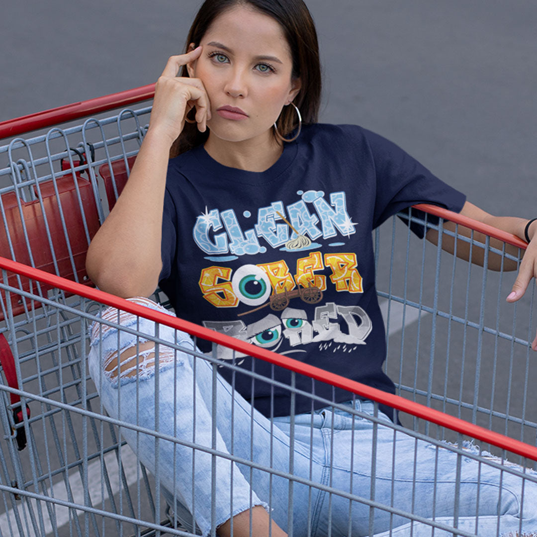 A woman sitting in a metal shopping cart wearing a navy blue graphic t-shirt with 'CLEAN SOBER GADGETS' printed on the front, paired with light-wash ripped jeans.