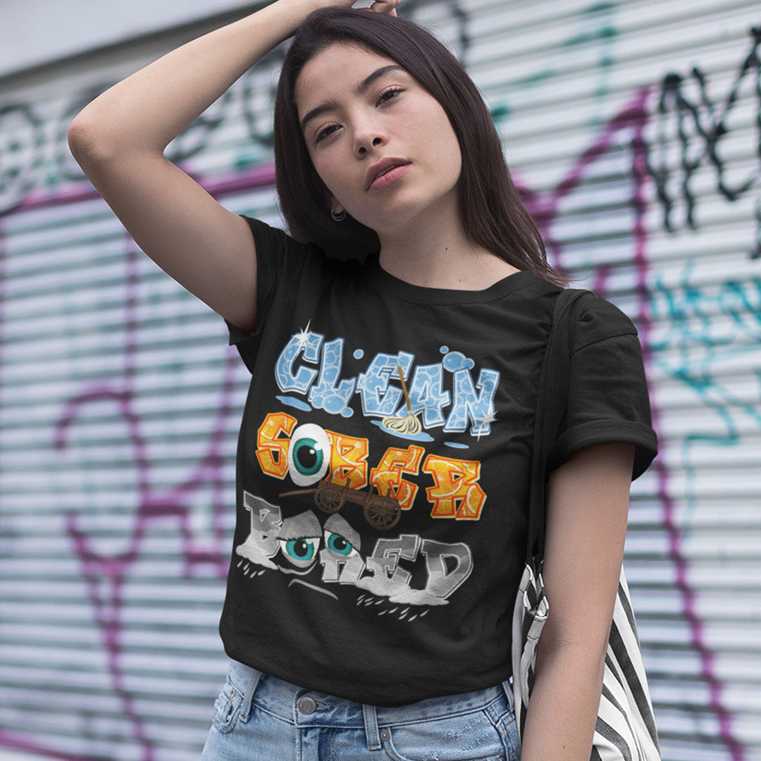 A woman wearing a black graphic t-shirt with the text 'CLEAN GOREY LORED' in stylized blue, orange, and white lettering, paired with light-wash denim jeans, posing in front of a graffiti-covered metal shutter.
