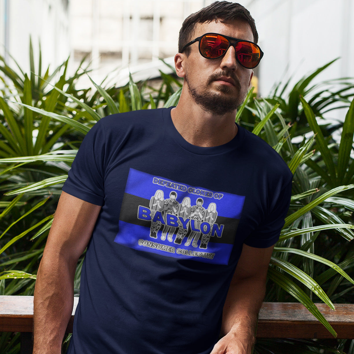 Man wearing a French navy blue t-shirt with a graphic design, sitting outdoors with plants in the background. This non conformist t shirt is for people with more than a pessimistic view of society and what we have become. This unique design has a Rastafarian hint as it suggests we fill our world with unnecessary distractions, ignoring more important issues. Made from 100% organic ring-spun cotton, this anti consumerism t shirt is unisex, high-quality, super comfy, and best of all, eco-friendly.