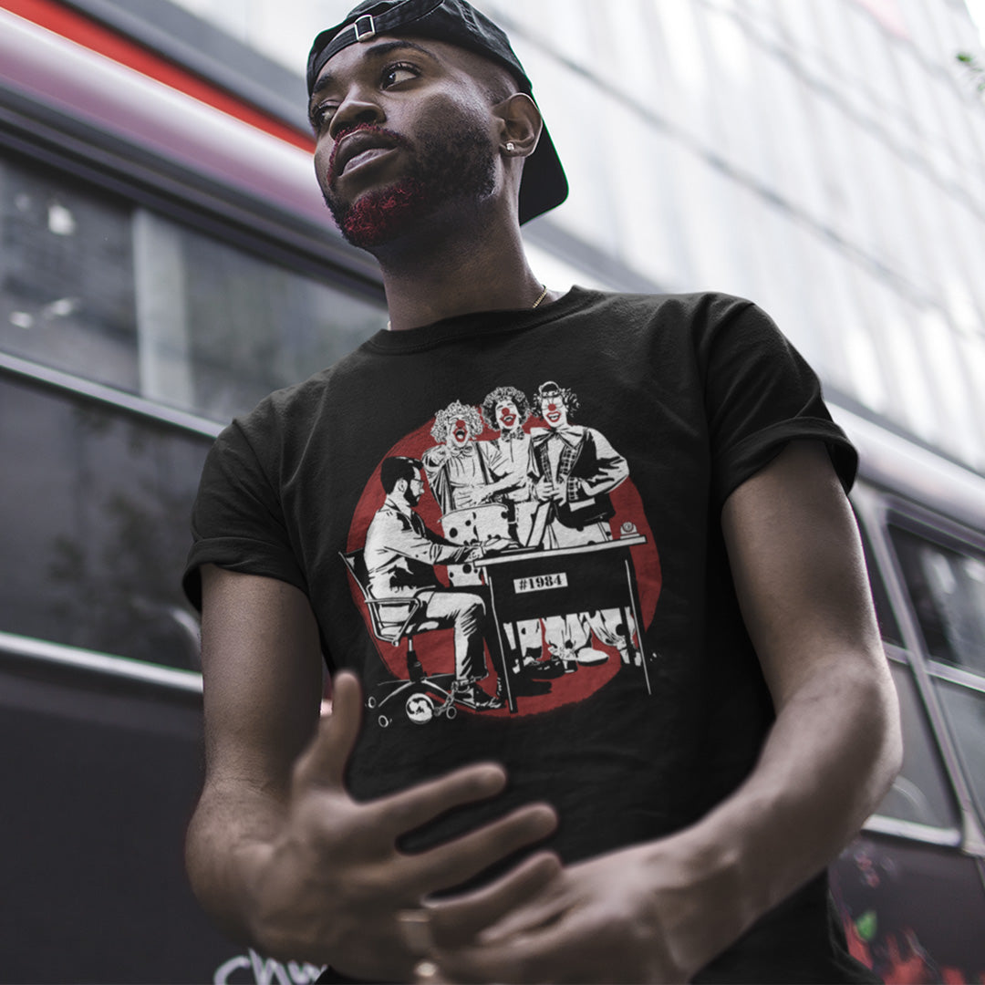 A man wearing a baseball cap backwards and a black short sleeved crew neck t-shirt with a white and red graphic of three clowns laughing and pointing at an office worker.