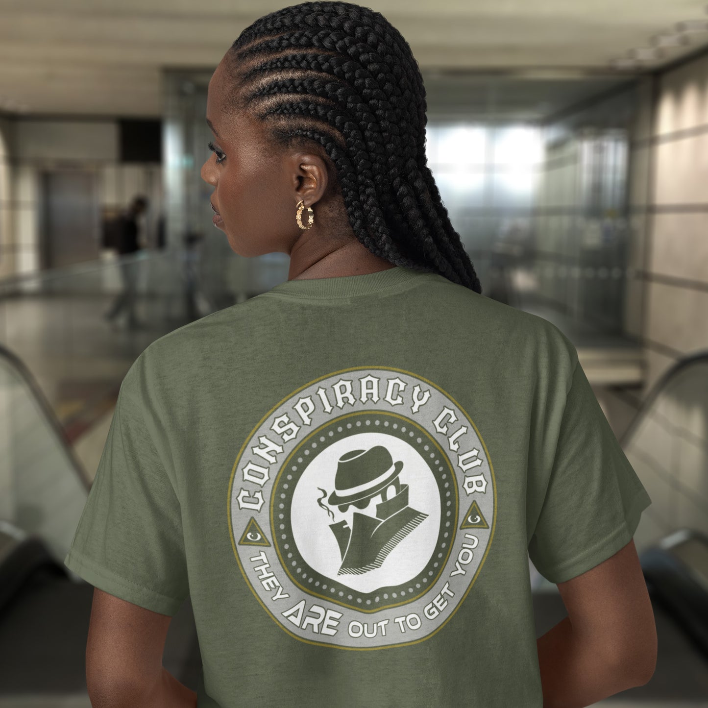 Person wearing a khaki t-shirt with 'Conspiracy Club' design in an indoor setting. A conspiracy theorist t shirt. These days there's a conspiracy theory for every occasion, join the club, expand your alternative imagination. Think outside the box. Underground art on apparel. Made from 100% organic ring-spun cotton, these conspiracy theory t shirts are unisex, high-quality, super comfy, and best of all, eco-friendly.