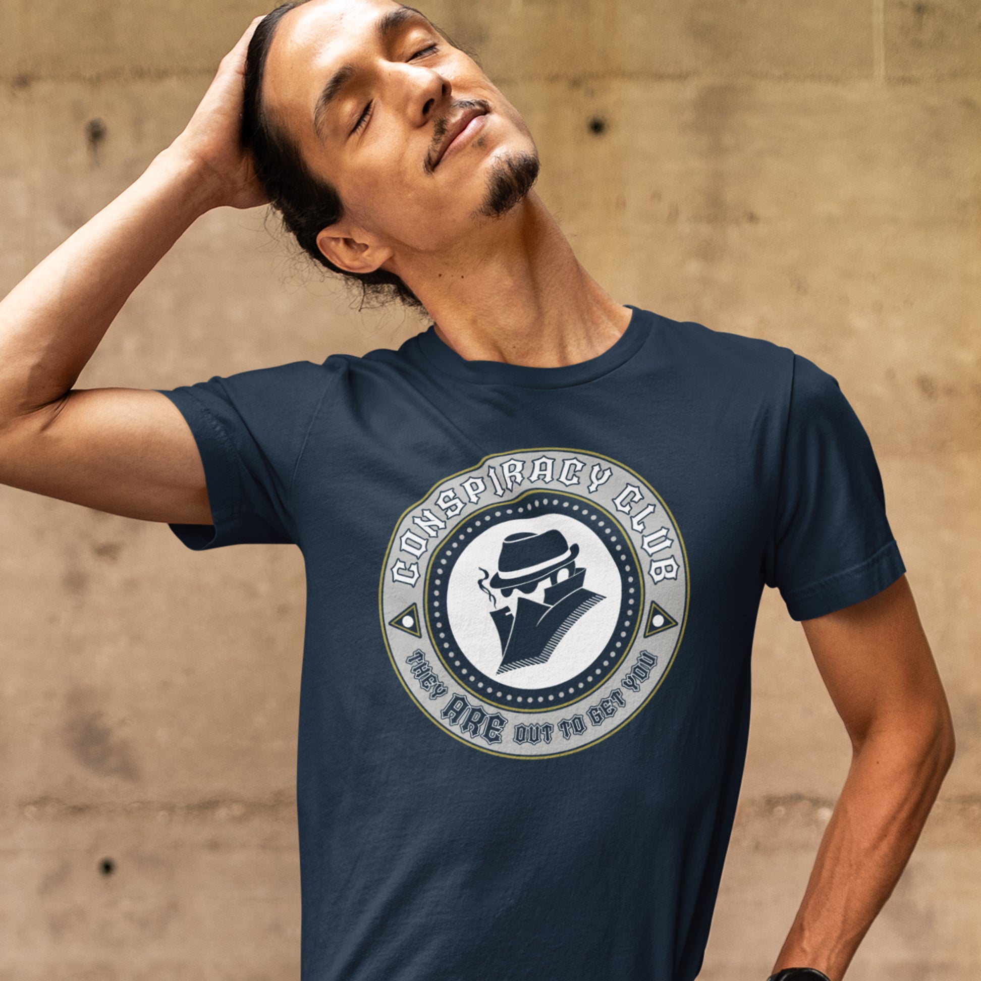 Man wearing a navy blue t-shirt with a 'Conspiracy Club' logo against a beige wall. These days there's a conspiracy theory for every occasion, join the club, expand your alternative imagination. Think outside the box. Underground art on apparel. Made from 100% organic ring-spun cotton, this conspiracy theory t-shirt is unisex, high-quality, super comfy, and best of all, eco-friendly.