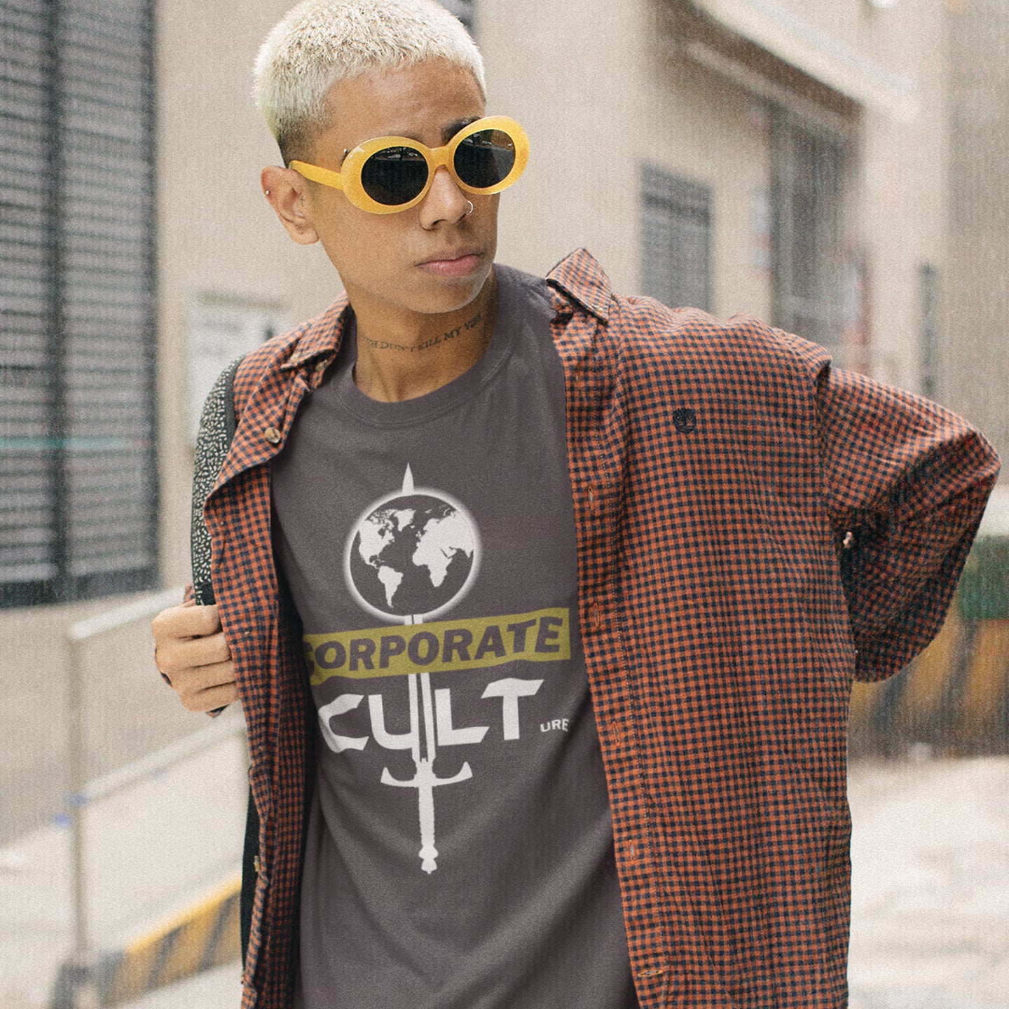 Person wearing an anthracite t-shirt with a graphic design and yellow sunglasses, standing outdoors. This anti corporate t shirt suggests the corporate mind set and dehumanisation of people is like that of a religious cult. Welcome to corporate culture, the enemy of the planet. Made from 100% organic ring-spun cotton, these social issues t shirts are unisex, high-quality and eco-friendly.