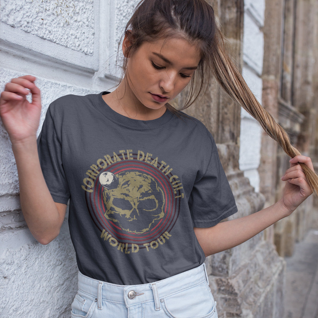 A woman looking down while playing with her hair, standing against a wall and wearing an anthracite  short sleeved crew neck t-shirt with a skull shaped dead planet graphic with "CORPORATE DEATH CULT WORLD TOUR" printed around it.