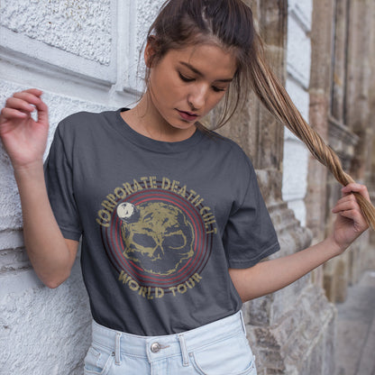 A woman looking down while playing with her hair, standing against a wall and wearing an anthracite  short sleeved crew neck t-shirt with a skull shaped dead planet graphic with "CORPORATE DEATH CULT WORLD TOUR" printed around it.