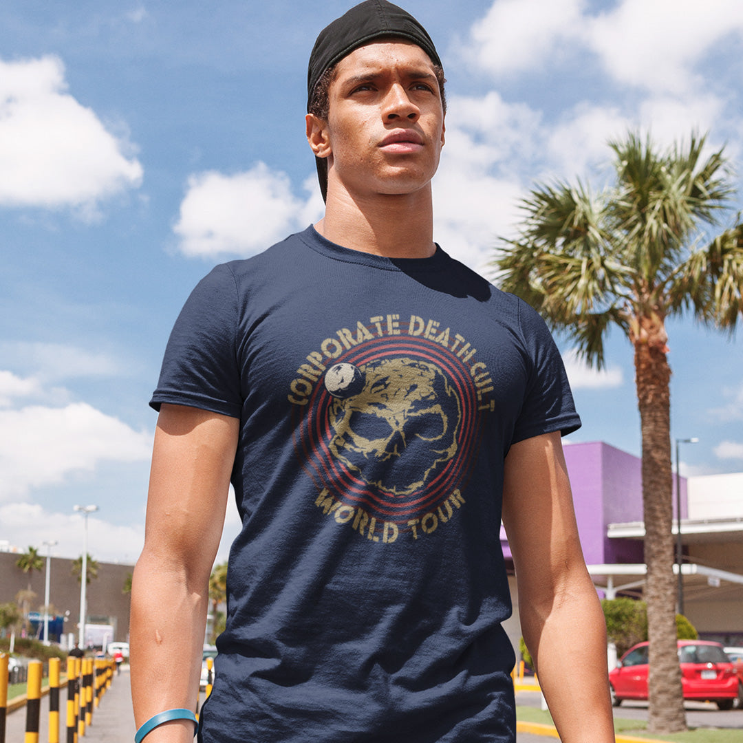 A man with a baseball cap on backwards, standing in the street and wearing a French navy blue  short sleeved crew neck t-shirt with a skull shaped dead planet graphic with "CORPORATE DEATH CULT WORLD TOUR" printed around it.