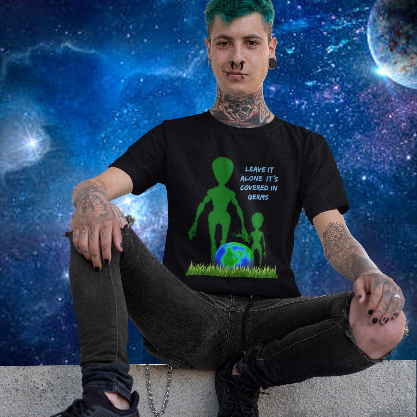 Person wearing a black t-shirt with an alien design and text, sitting against a space-themed background. A funny alien t-shirt design with a subtle overpopulation message. In the grand scheme of things, our planet and universe could just be the school project of a child from a superior race. Made from 100% organic ring-spun cotton, this witty alien t-shirt is unisex, high-quality, super comfy, and best of all, eco-friendly.