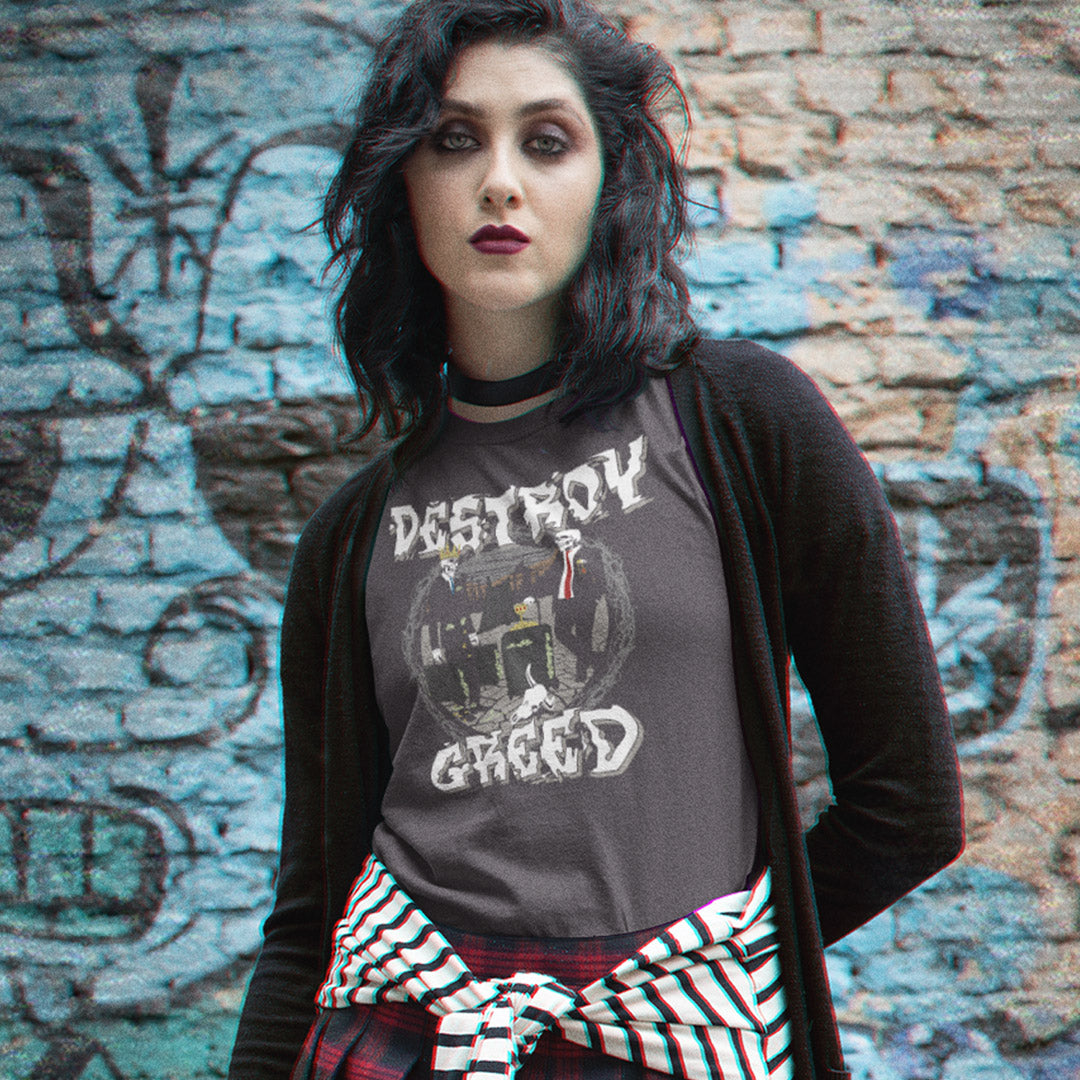 A woman with dark hair stands in front of a graffiti-covered brick wall, wearing a dark gray graphic t-shirt with the words 'DESTRUCTION GREED' and a black cardigan, with a red, black, and white striped shirt tied around her waist.