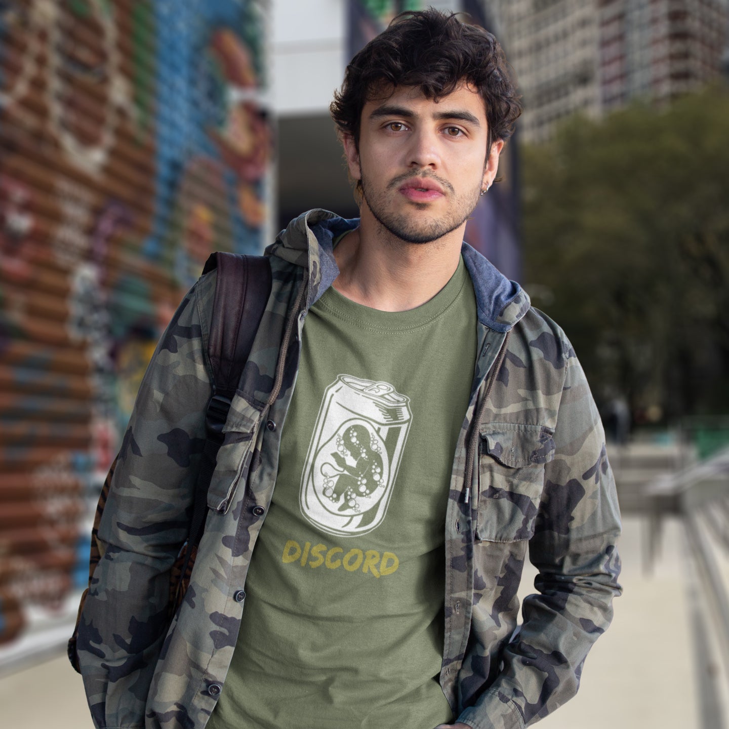 Man wearing a khaki t-shirt with Discord logo and text, standing in an urban setting. A Banksy style t-shirt suggesting a discord between modern generations and nature. A stencil art testament to the soda pop generation. Brought up on energy drinks and born with a Wi-Fi connection. Made from 100% organic ring-spun cotton, this street art t-shirt is unisex, high-quality, super comfy, and best of all, eco-friendly.