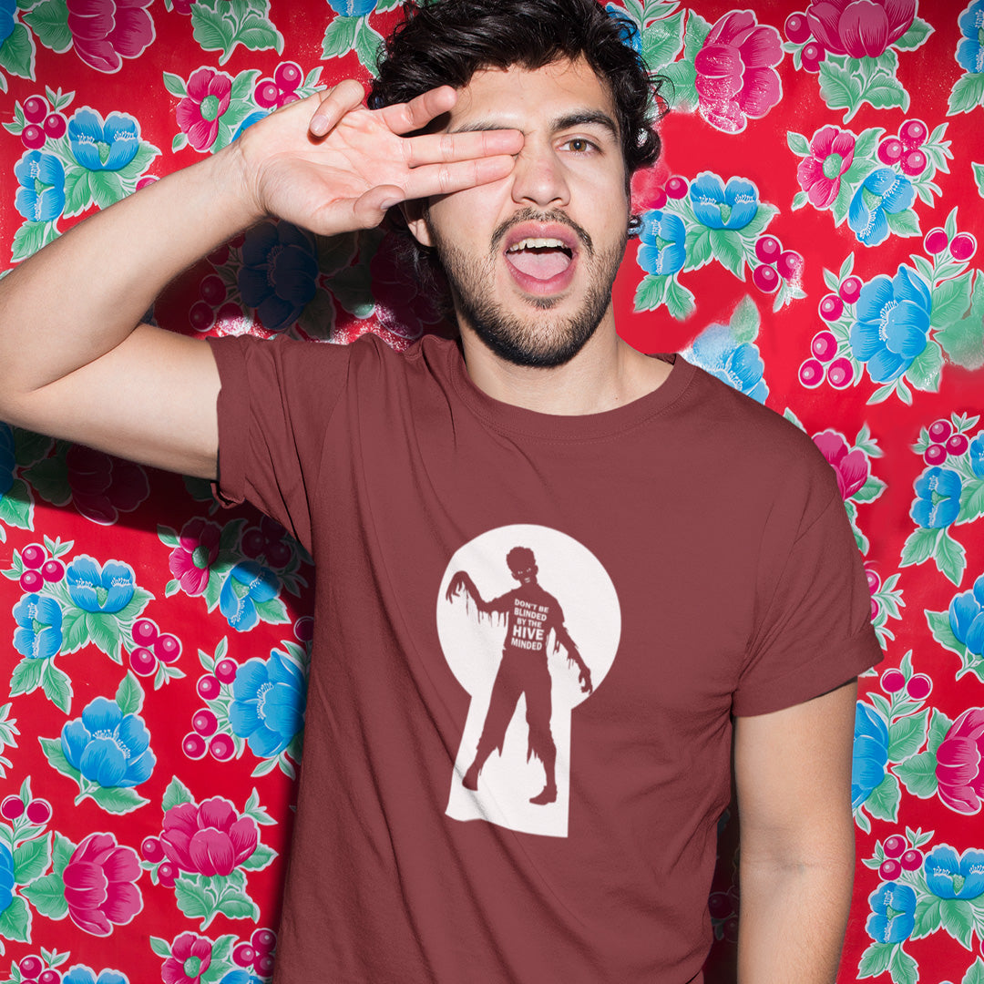 A man with two fingers over his eye, standing in front of flower wallpaper, wearing a burgundy  t-shirt with a zombie through a key hole design with "DON'T BE BLINDED BY THE HIVE MINDED" text.