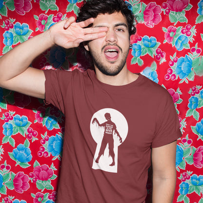 A man with two fingers over his eye, standing in front of flower wallpaper, wearing a burgundy  t-shirt with a zombie through a key hole design with "DON'T BE BLINDED BY THE HIVE MINDED" text.