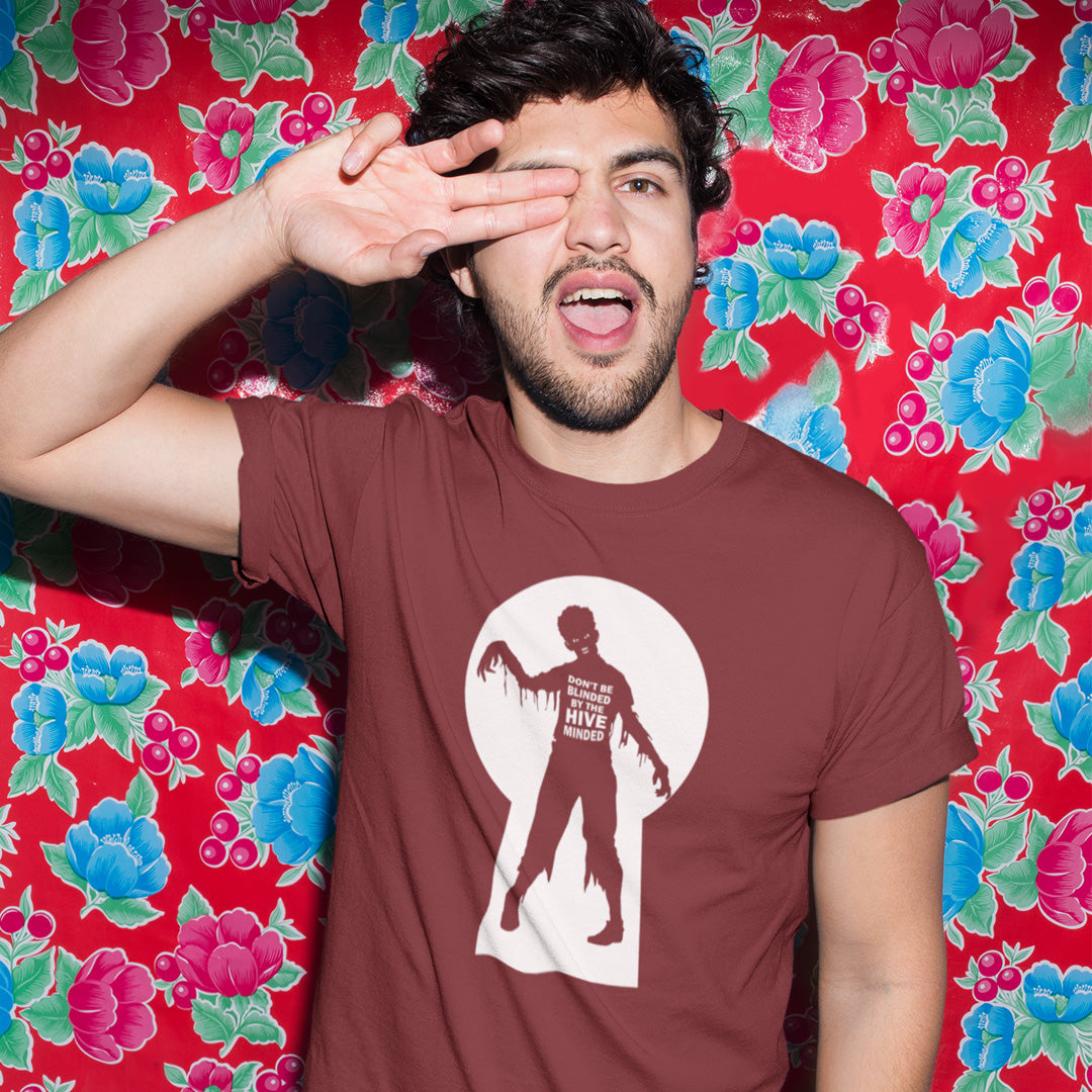 A man wearing a brown t-shirt with a zombie graphic, posing with his hand over his eye in front of a red floral backdrop.