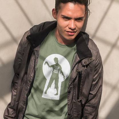 A man wearing a dark gray windbreaker jacket over an olive green graphic t-shirt with a zombie print, posing against a wall with window shadow patterns.