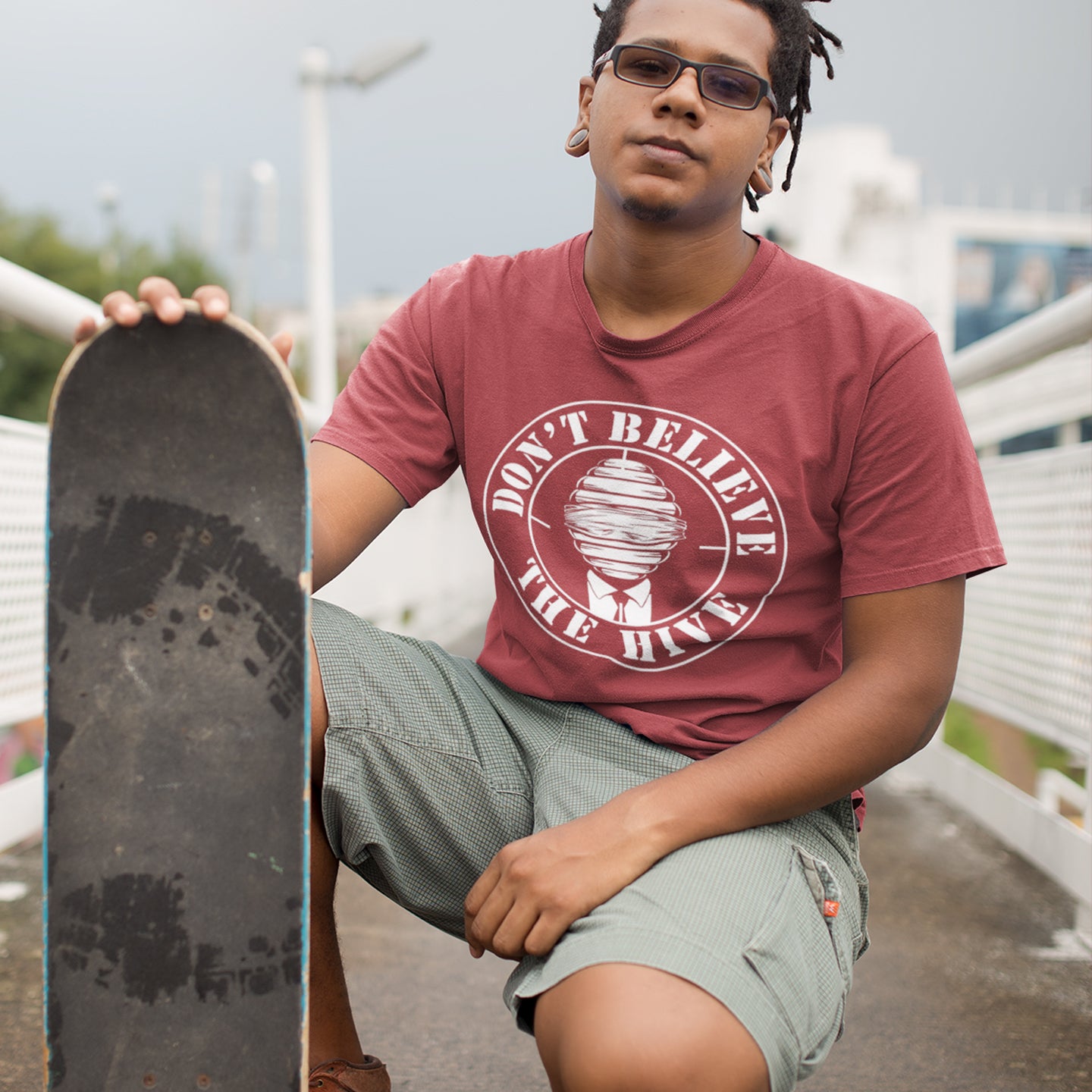 Person wearing a burgundy t-shirt with a graphic design, holding a skateboard outdoors. A street art t shirt design for an ever increasing world of media influence and misinformation. Can 'The Majority' be trusted any more ? Don't get blinded by the hive minded. Support independent thought with this unique urban art t shirt. Made from 100% organic ring-spun cotton, this social awareness t shirt is unisex, high-quality, super comfy, and best of all, eco-friendly.