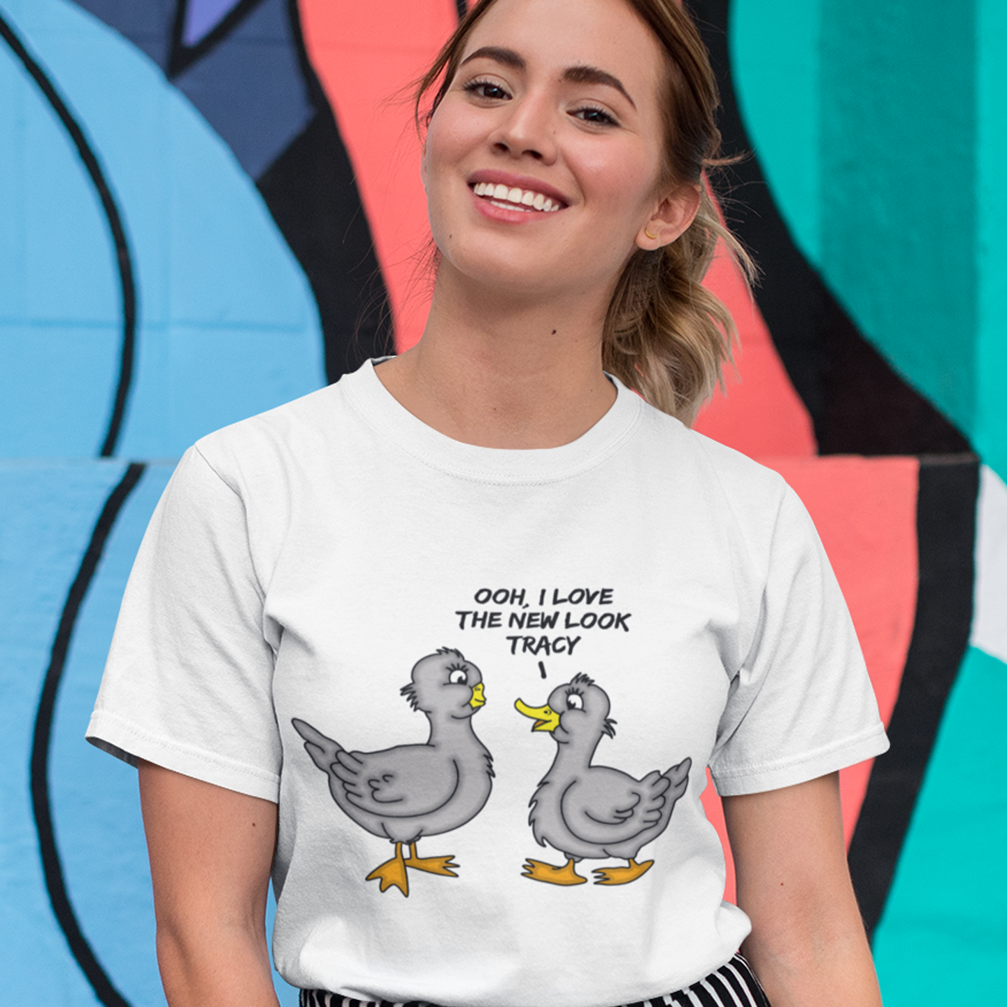 Person wearing a white t-shirt with cartoon ducks and text, standing against a colourful abstract background. A social issues t shirt design mocking the idea of lip fillers. In a alternative reality are ducks having surgery to make their lips look like humans ? Counterculture cartoon. Made from 100% organic ring-spun cotton, this social awareness t shirt is unisex, high-quality, super comfy, and best of all, eco-friendly.