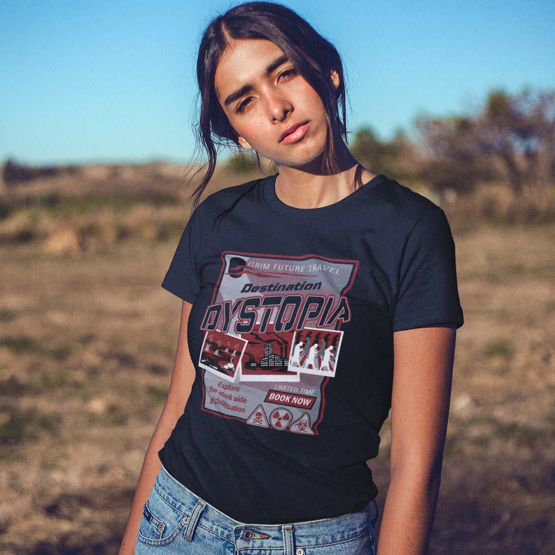 A woman standing outdoors, wearing a French navy blue , short sleeved, crew necked t-shirt with a spoof travel poster design with "Destination Dystopia" text.