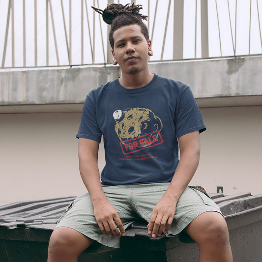 A man with a dreadlock sits on a dark metal dumpster outdoors, wearing a French navy blue graphic t-shirt featuring a graphic of a brown textured planet with a small white moon, overlaid with a red 'FOR SALE' stamp and red text below reading 'ONLY 8,000,000,000. PREVIOUS CARELESS OWNERS.