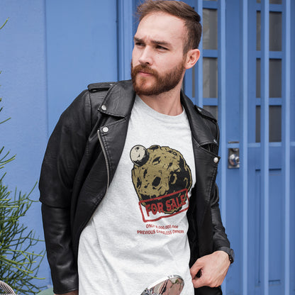 A man wearing a light grey graphic t-shirt featuring a graphic of a brown textured planet with a small white moon, overlaid with a red 'FOR SALE' stamp and red text below reading 'ONLY 8,000,000,000. PREVIOUS CARELESS OWNERS.