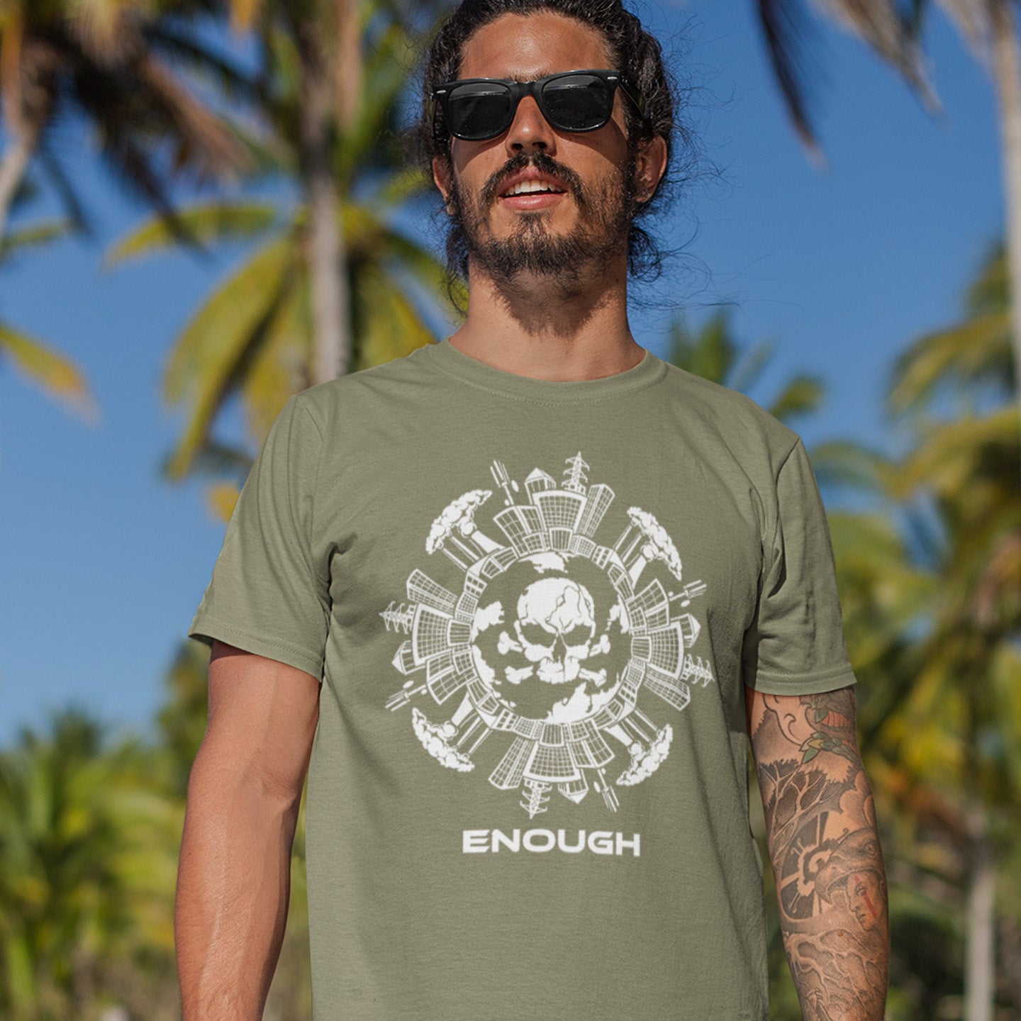 Man wearing a khaki t-shirt with a skull  design and 'ENOUGH' text, standing outdoors with palm trees in the background. A save the world t shirt highlighting an overcrowding concern. This original design is for all nature lovers who think that enough is enough and don't want to live in a concrete jungle. Made from 100% organic ring-spun cotton, this save the earth t shirt is unisex, high-quality, super comfy, and best of all, eco-friendly.