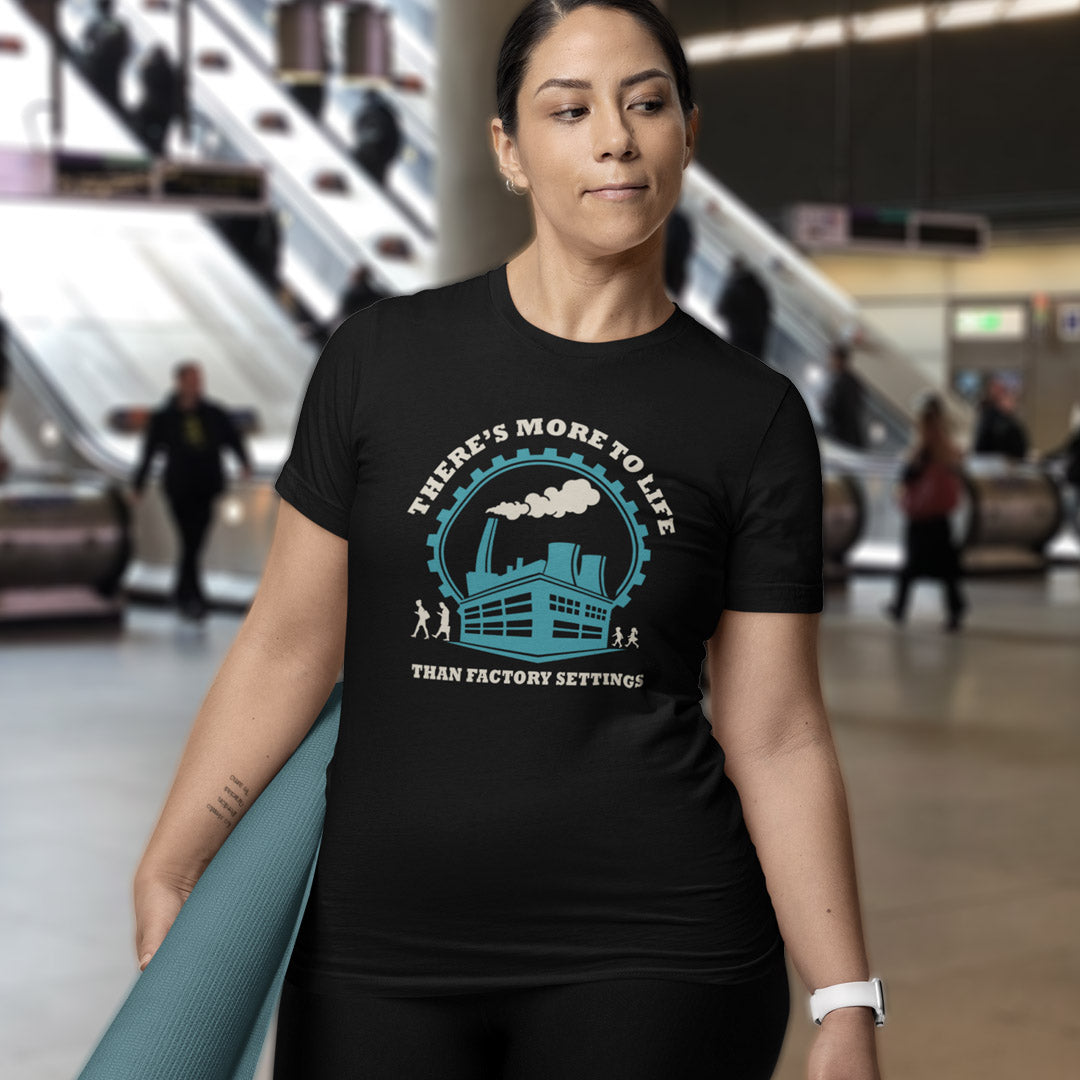 A woman wearing a black graphic t-shirt with a blue and white factory illustration and the text 'THERE'S MORE TO LIFE THAN FACTORY SETTINGS'. She is holding a teal yoga mat in a blurred indoor environment with escalators in the background.
