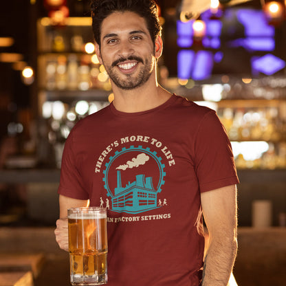 A smiling man in a bar setting wearing a burgundy t-shirt with a teal and white graphic that reads 'THERE'S MORE TO LIFE THAN FACTORY SETTINGS'. He is holding a large glass mug filled with an amber-coloured drink.
