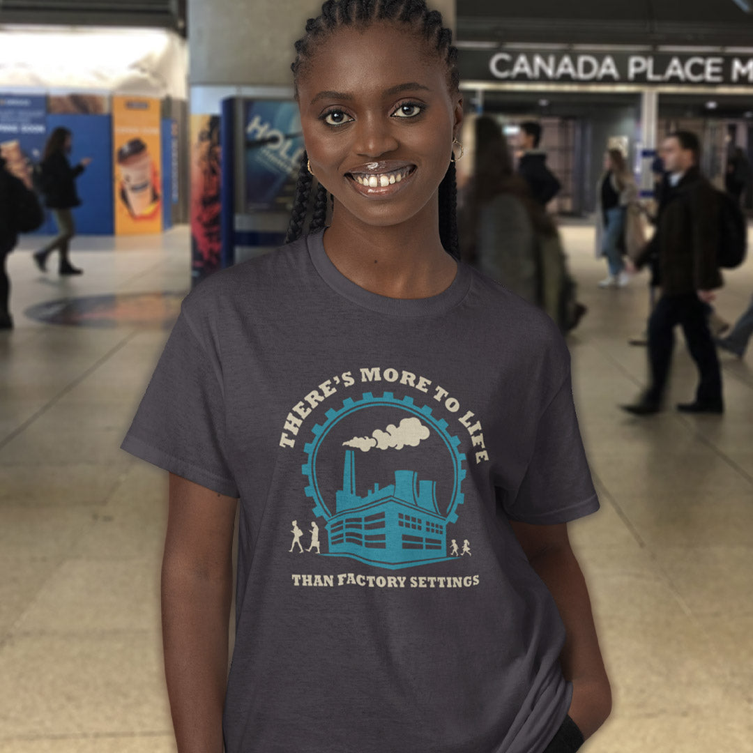 A smiling woman wearing an anthracite graphic t-shirt in a busy indoor public space. The t-shirt features a light blue and cream graphic of a factory with the text 'THERE'S MORE TO LIFE THAN FACTORY SETTINGS'.