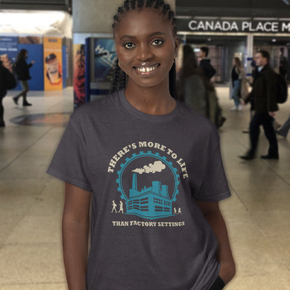 A smiling woman wearing an anthracite graphic t-shirt in a busy indoor public space. The t-shirt features a light blue and cream graphic of a factory with the text 'THERE'S MORE TO LIFE THAN FACTORY SETTINGS'.