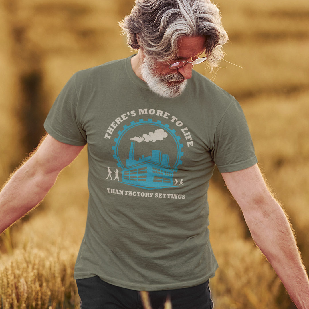 A man wearing an khaki graphic t-shirt with a blue and white industrial factory design and the text 'THERE'S MORE TO LIFE THAN FACTORY SETTINGS', standing in a golden wheat field.