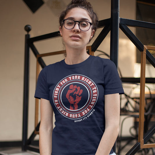 A woman wearing a navy blue graphic t-shirt with a red and white fist design. She is wearing black-rimmed glasses and standing in front of a black metal staircase.