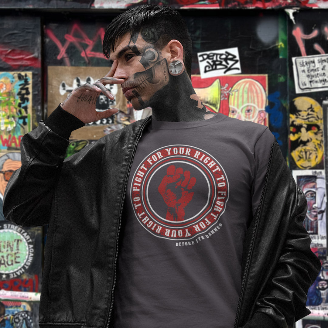 A man with blackface makeup and a mechanical eye wears a dark gray graphic t-shirt and a black leather jacket, standing in front of a graffiti-covered wall.