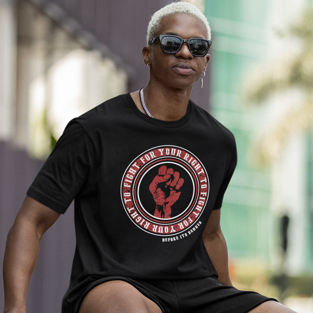 A man wearing a black graphic t-shirt with a red fist print and the text 'TO FIGHT FOR YOUR RIGHT TO FIGHT FOR YOUR RIGHT' on the front, set against a blurred outdoor background.