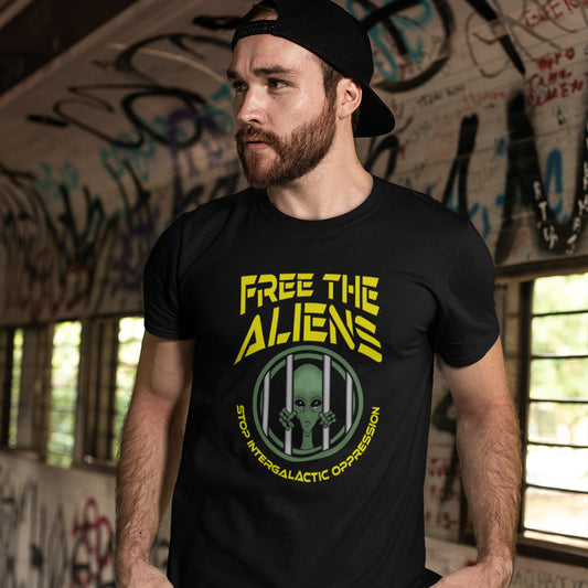 A man wearing a black t-shirt with a yellow and green graphic that reads 'FREE THE ALIENS' and 'STOP INTERSALTIC OPPRESSION' with an alien in a cage illustration. He is wearing a black baseball cap and standing in a graffiti-covered indoor space.