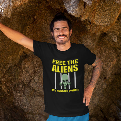 Man wearing a black t-shirt with 'Free the Aliens' text and graphic, standing against a rocky background. This funny alien t shirt alludes to there being more than just UFO crashes that we are not being told about. With all these alien sightings they must detaining them somewhere ! Made from 100% organic ring-spun cotton, this conspiracy theory t shirt is unisex, high-quality, super comfy, and best of all, eco-friendly.