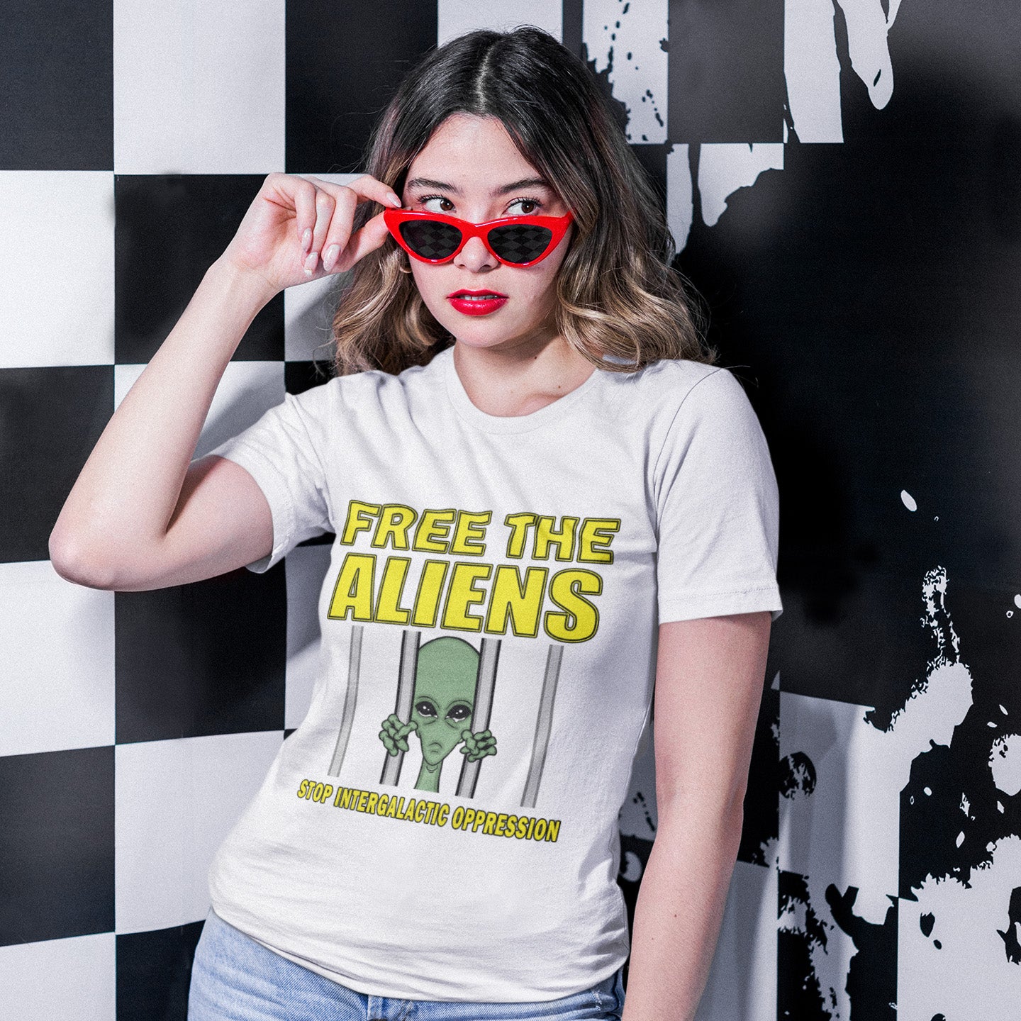 Woman wearing a white t-shirt with 'Free the Aliens' graphic design, standing against a black and white chequered wall. This funny alien t shirt alludes to there being more than just UFO crashes that we are not being told about. With all these alien sightings they must detaining them somewhere ! Made from 100% organic ring-spun cotton, this conspiracy theory t shirt is unisex, high-quality, super comfy, and best of all, eco-friendly.