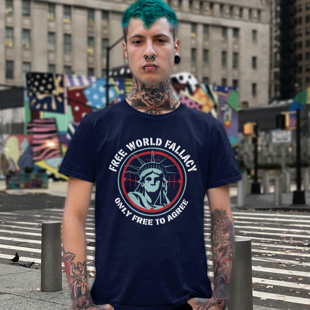 A man with teal hair, tattoos, and a nose ring stands on a city crosswalk wearing a navy blue graphic t-shirt. The shirt features a circular design with the Statue of Liberty and the text 'FREE WORLD FALLACY' and 'ONLY FREE TO AGREE'.