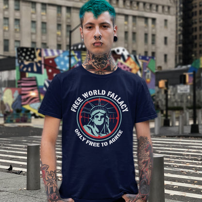 A man with teal hair, tattoos, and a nose ring stands on a city crosswalk wearing a navy blue graphic t-shirt. The shirt features a circular design with the Statue of Liberty and the text 'FREE WORLD FALLACY' and 'ONLY FREE TO AGREE'.