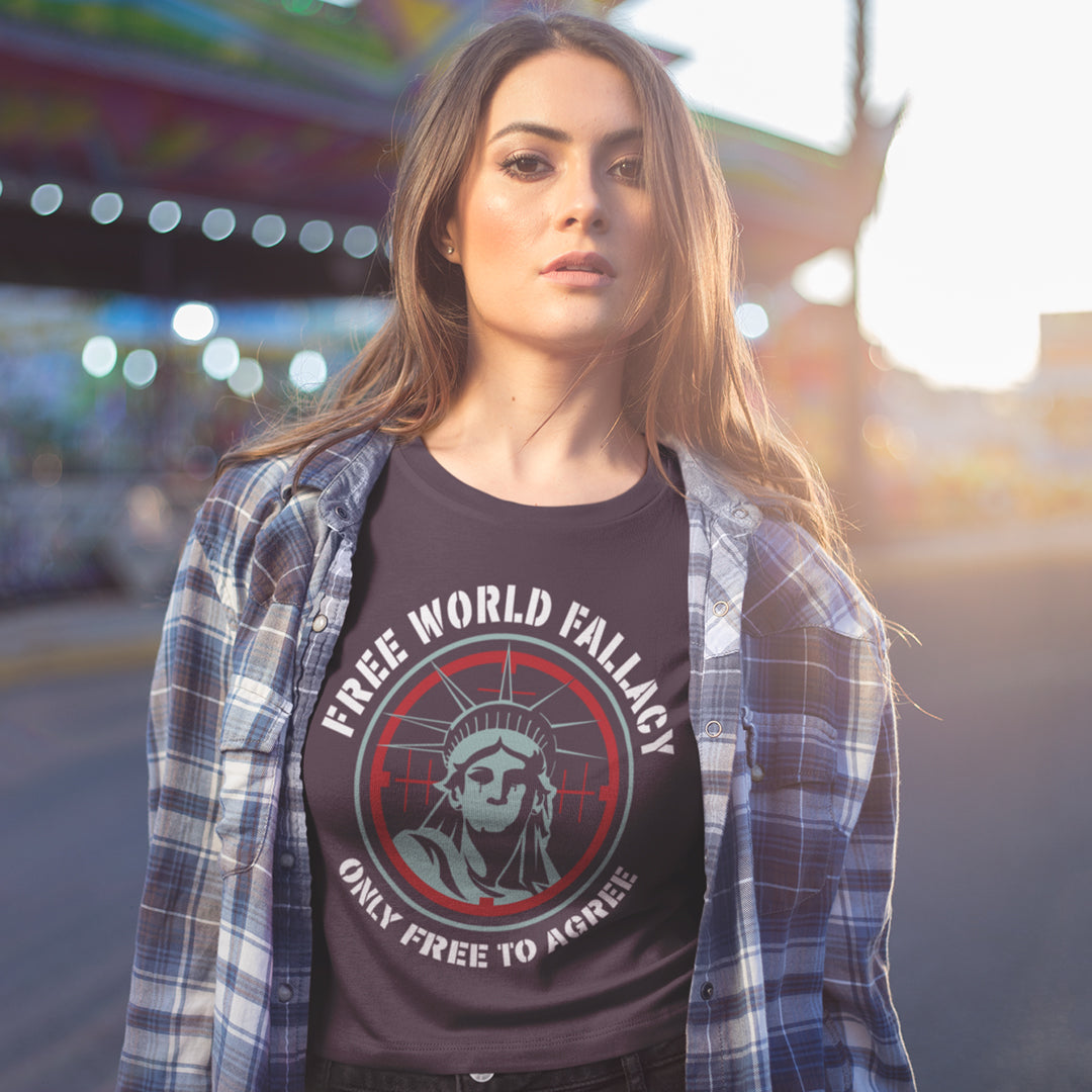 A woman wearing a dark graphic t-shirt with a plaid shirt open over it, standing outdoors in a blurred urban environment. The t-shirt features a circular graphic of the Statue of Liberty with the text 'FREE WORLD FALLACY' and 'ONLY FREE TO AGREE'.