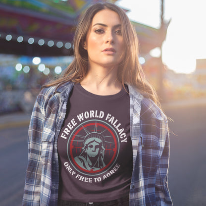 A woman wearing a dark graphic t-shirt with a plaid shirt open over it, standing outdoors in a blurred urban environment. The t-shirt features a circular graphic of the Statue of Liberty with the text 'FREE WORLD FALLACY' and 'ONLY FREE TO AGREE'.