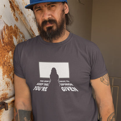 A man wearing a dark gray graphic t-shirt and a blue cap, standing in front of a rusty metal door. The t-shirt features a white graphic with a silhouette and the text 'YOU DON'T HAVE TO KEEP THE OPINION YOU'RE GIVEN'.