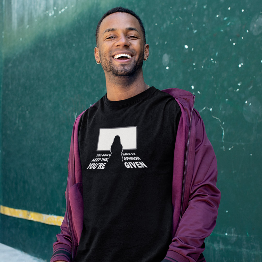 A smiling man wearing a black graphic t-shirt and a purple zip-up hoodie, standing against a textured green wall.