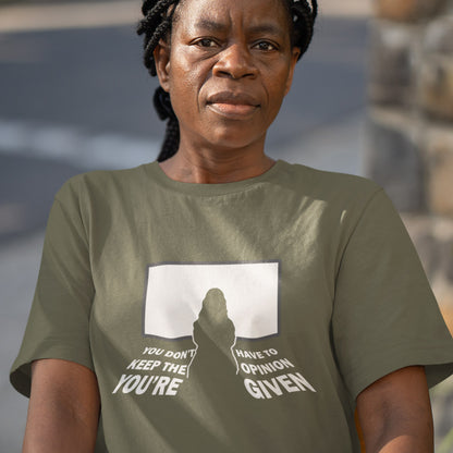 A woman wearing an olive green graphic t-shirt with a white print that reads 'YOU DON'T HAVE TO KEEP THE OPINION YOU'RE GIVING' alongside a silhouette graphic, set against a blurred outdoor background.