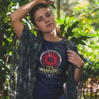 A woman wearing a navy blue graphic t-shirt with a circular red, yellow, and white 'The Ancient Art Good Manners' print, styled with a sheer black patterned kimono over a lush green forest background.