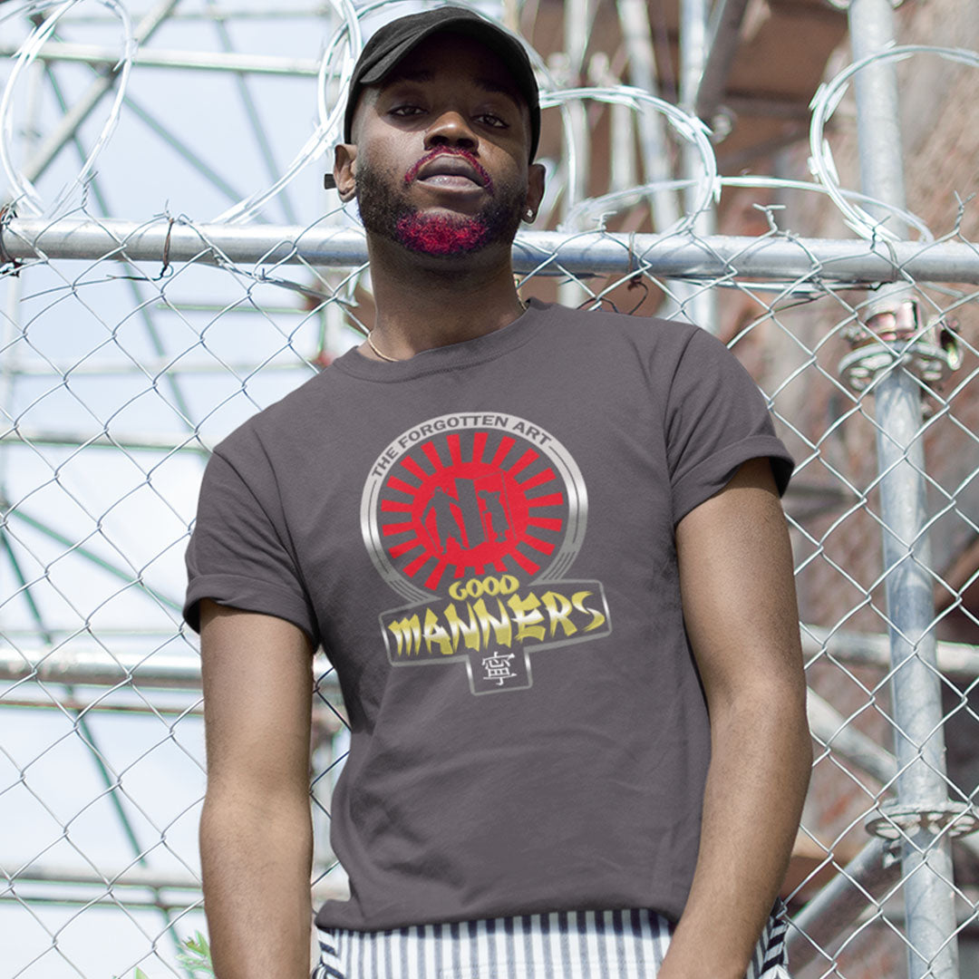 A man wearing a dark gray t-shirt with a red and yellow 'Good Manners' graphic, standing in front of a chain-link fence.