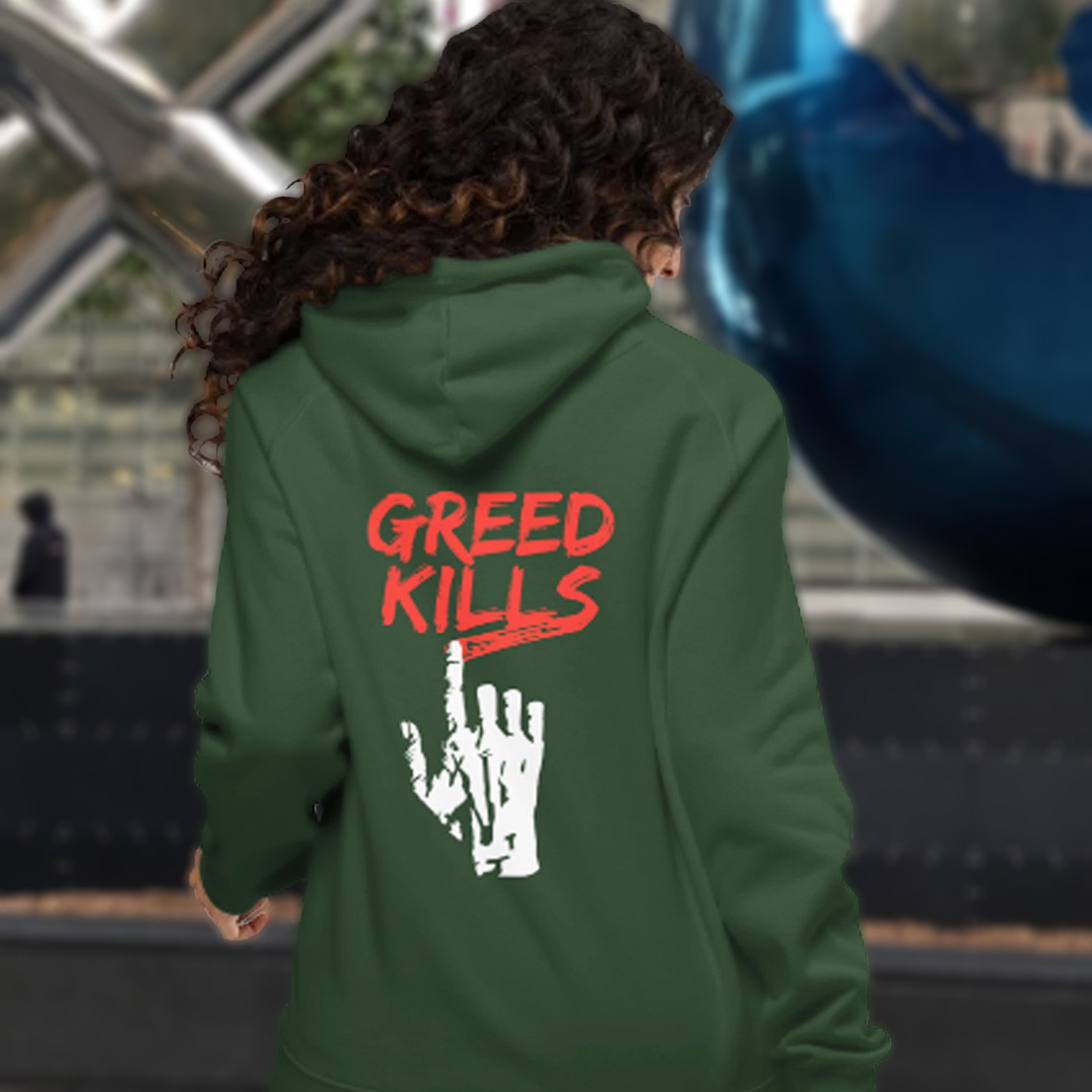 Person wearing a green hoodie with 'GREED KILLS' text on the back, standing in an outdoor setting. An anti-capitalism t-shirt focusing on greed. It's the slow-creeping, strong-holding virus that has been with us since the beginning of time, yet we still can't find a cure. Comfortable and practical. It's extra soft and has a convenient front pouch pocket. Made from organic cotton and recycled polyester, this social awareness hoodie is a great eco-friendly choice for the anti-capitalist.