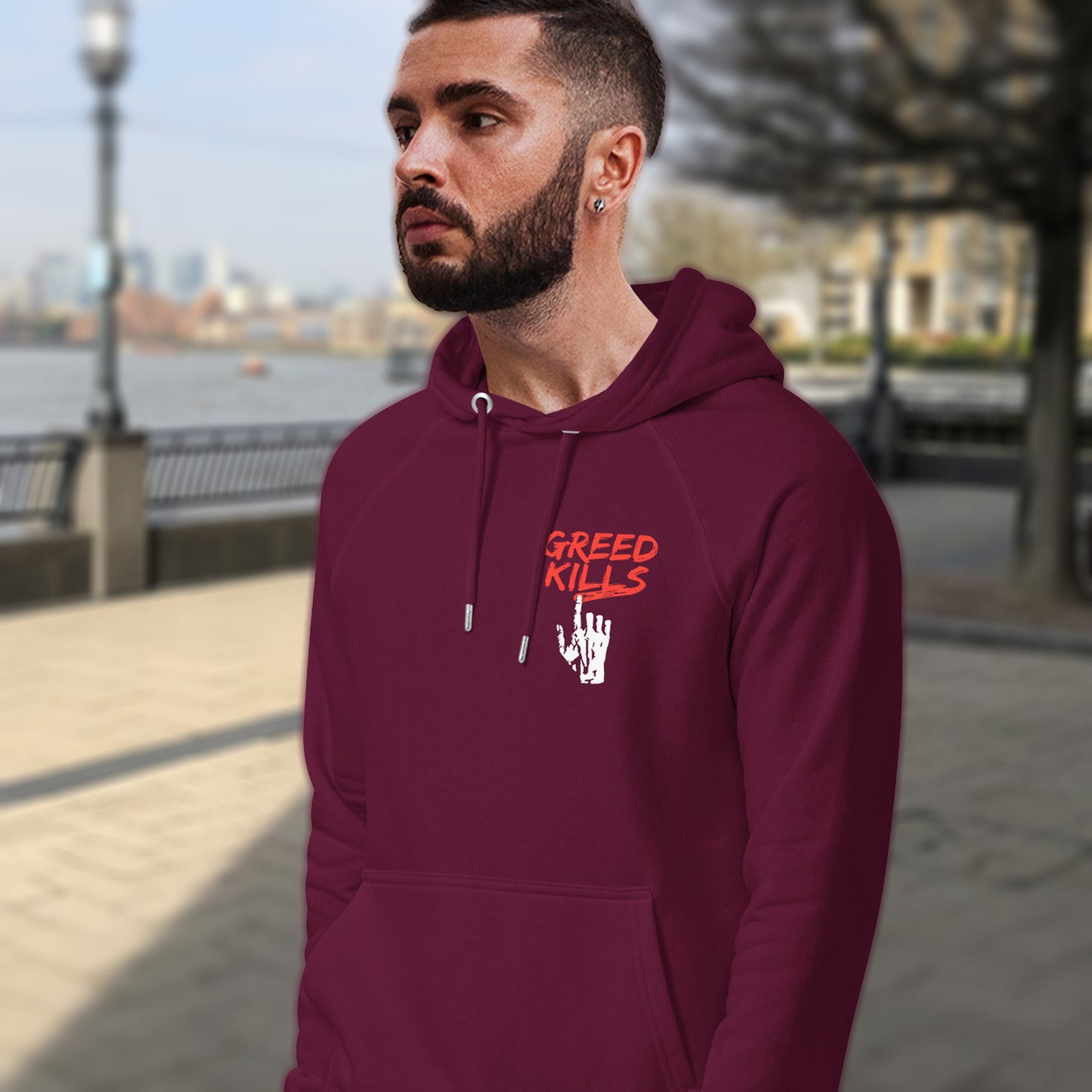Man wearing a maroon hoodie with 'Greed Kills' text outdoors. An anti-capitalism t-shirt focusing on greed. It's the slow-creeping, strong-holding virus that has been with us since the beginning of time, yet we still can't find a cure. Comfortable and practical. It's extra soft and has a convenient front pouch pocket. Made from organic cotton and recycled polyester, this social awareness hoodie is a great eco-friendly choice for the anti-capitalist.