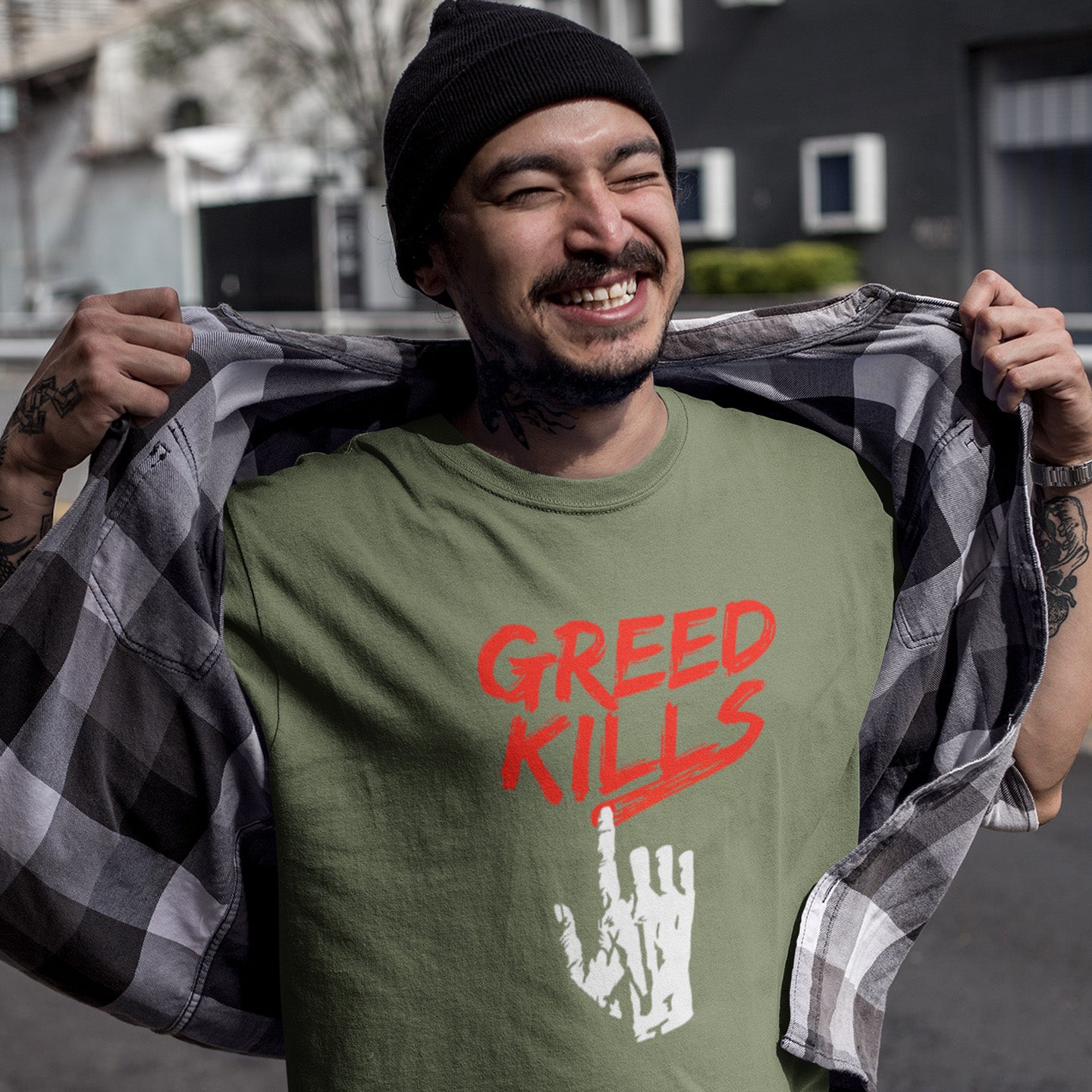 Person wearing a khaki t-shirt with 'Greed Kills' text and middle finger gesture, holding a chequered shirt. This anti capitalism t shirt opposes greed, the slow-creeping, strong-holding virus that has been with us since the beginning of time, yet we still can't find a cure. A morality t-shirt design promoting social awareness of the effect of greed on the world. Made from 100% organic ring-spun cotton, this ant capitalist t shirt is unisex, high-quality, super comfy, and best of all, eco-friendly.