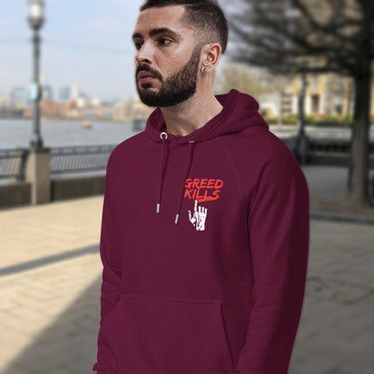 Man wearing a  burgundy hoodie with 'Greed Kills' text outdoors. An anti-capitalism hoodie design focusing on greed. It's the slow-creeping, strong-holding virus that has been with us since the beginning of time, yet we still can't find a cure. Comfortable and practical. It's extra soft and has a convenient front pouch pocket. Made from organic cotton and recycled polyester, this non conformist hoodie is a great eco-friendly choice for the anti-capitalist.