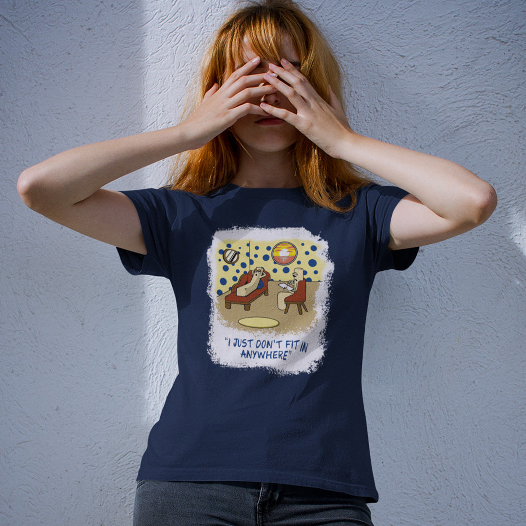 A woman with red hair stands against a textured light grey wall, holding her hands over her eyes. She is wearing a French navy blue graphic t-shirt. The graphic features a cartoon illustration of a person sitting on a lounge chair in a theorists room , with a white distressed border and the text 'I JUST DON'T FIT IN ANYWHERE' below.