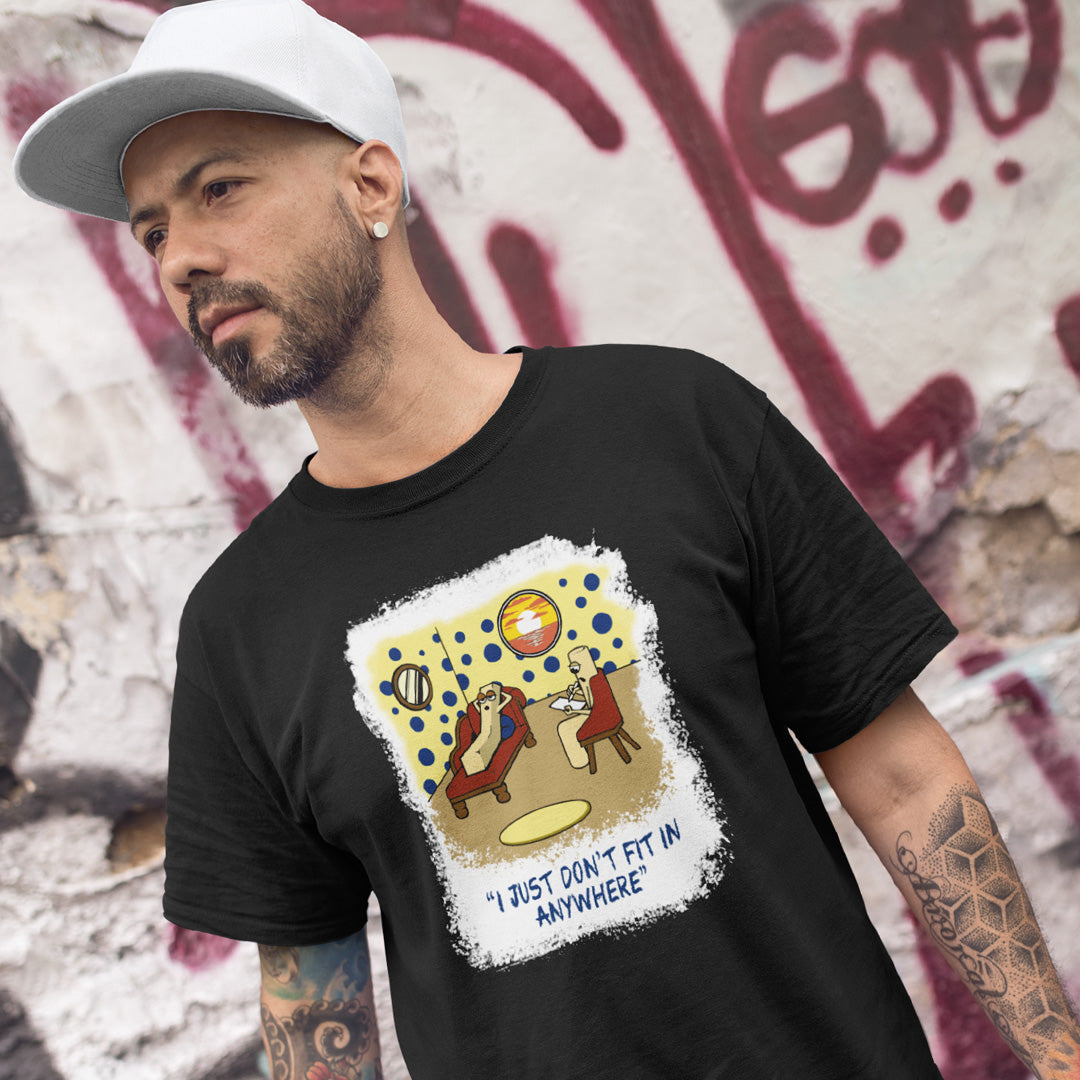 A man wearing a black graphic t-shirt with a retro-style illustration and the text 'I JUST DON'T FIT IN ANYWHERE' on the front, paired with a white baseball cap, posed against a graffiti-covered wall.